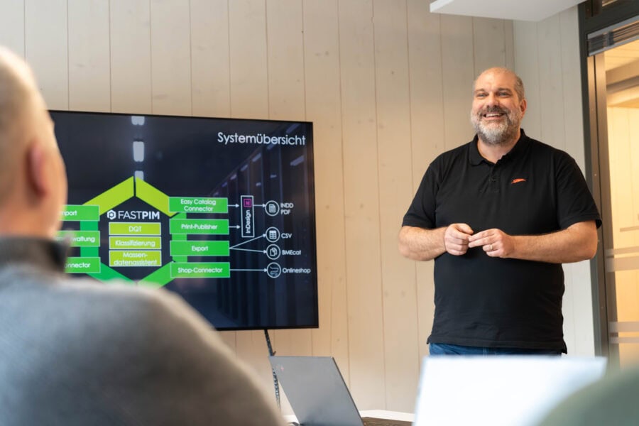Ein Mann steht vor einem Bildschirm und erklärt ein Systemübersichts-Diagramm zum Thema FASTPIM in einem Meetingraum, während zwei Personen im Vordergrund zuhören.
