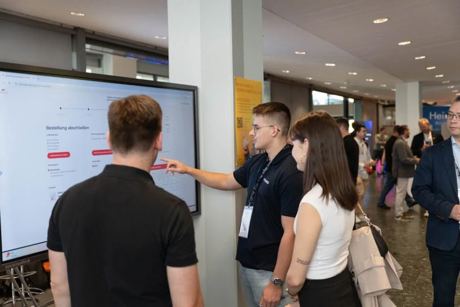IANEO-Mitarbeiter zeigt zwei Messebesuchern am Stand der IT.CON einen Shopware-Checkout „Bestellung abschließen" auf einem großen Bildschirm
